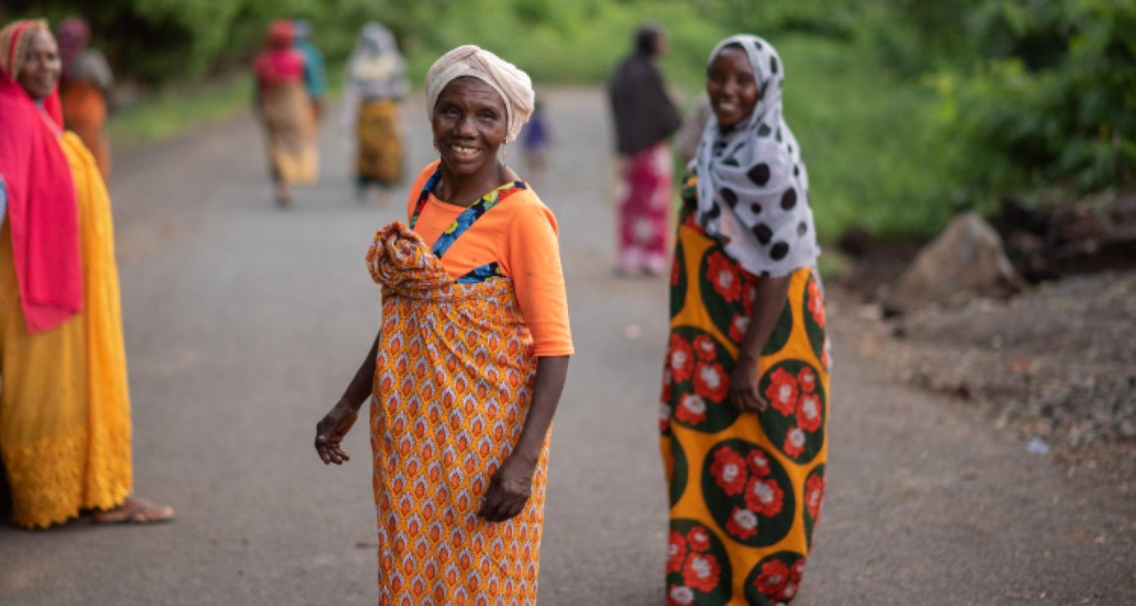Two woman in orange patterned dresses stand in the road turning to smile at the camera 