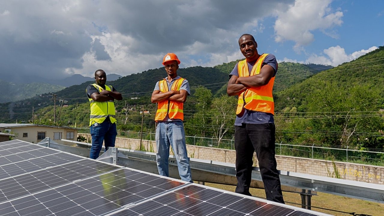 three men in hard hats and vests stand by solar panel 