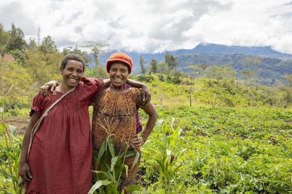 Two smiling women stand arm in arm in a green field with hills in the background.