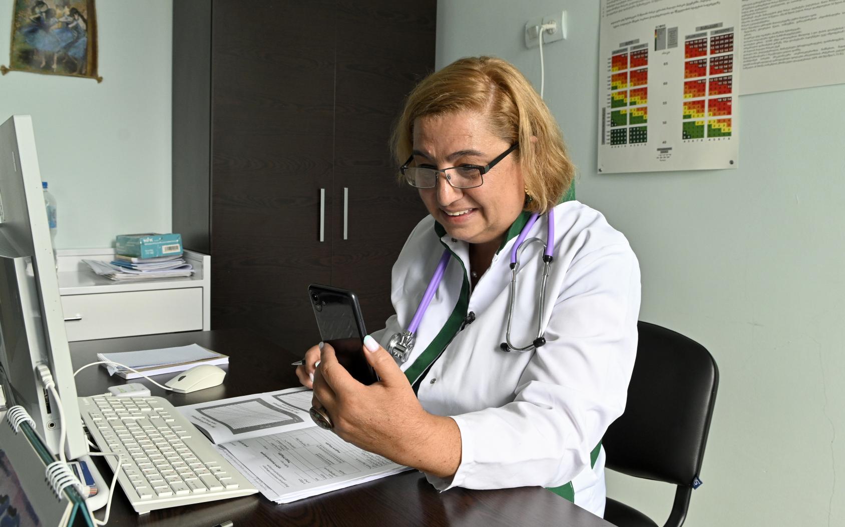 A woman in a white doctor's coat and stethoscope holds up a mobile phone as she sits at a desk in a clinic.