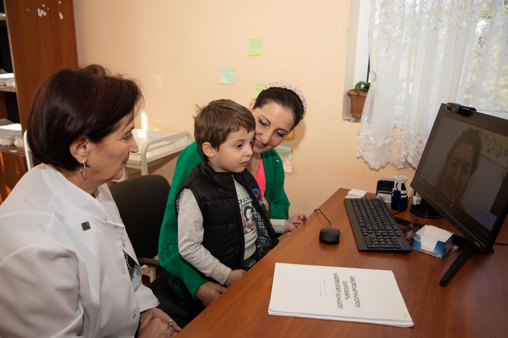 A little boy sits on the lap of a woman in a green dress, next to another woman in white clothing. He is talking to a doctor in a computer screen on a brown desk.