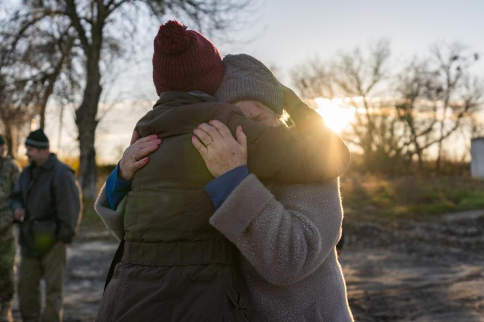 The image shows two individuals embracing each other outdoors during a sunset. They are surrounded by barren trees and bathed in the warm glow of the setting sun.