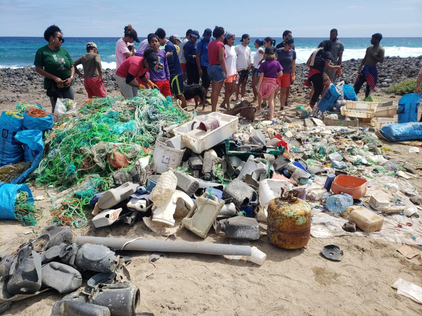 A group of people standing on a beach behind a large collection of assorted debris and trash. The waste includes items such as plastic bottles, nets, containers, a tire, and other miscellaneous items that have been washed up or left on the shore. This image highlights the environmental issue of ocean pollution and its impact on natural habitats.