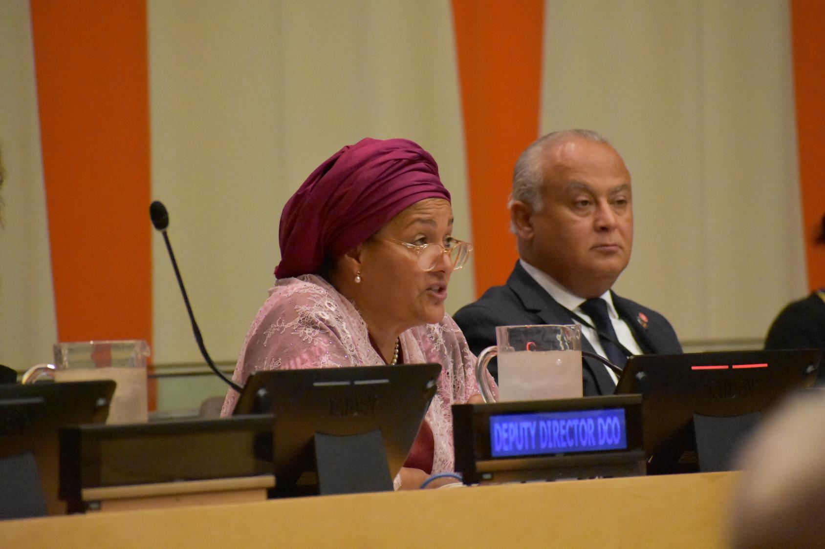 Image of two individuals seated at a conference. They are engaged in a discussion, with microphones and name tags on the table. The setting includes a backdrop of vertical orange and white panels.