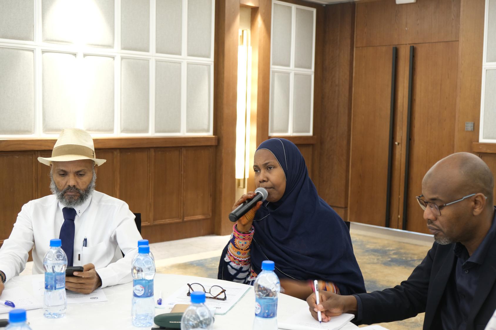 A woman in a head scarf is using a microphone at a table. Beside her is a man in a black suit and a man in a white suit wearing a hat 
