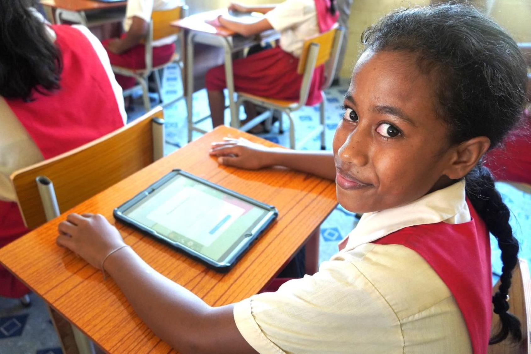 A student in a red uniform shirt with a white collar is seated at a desk in a classroom, using a tablet. 