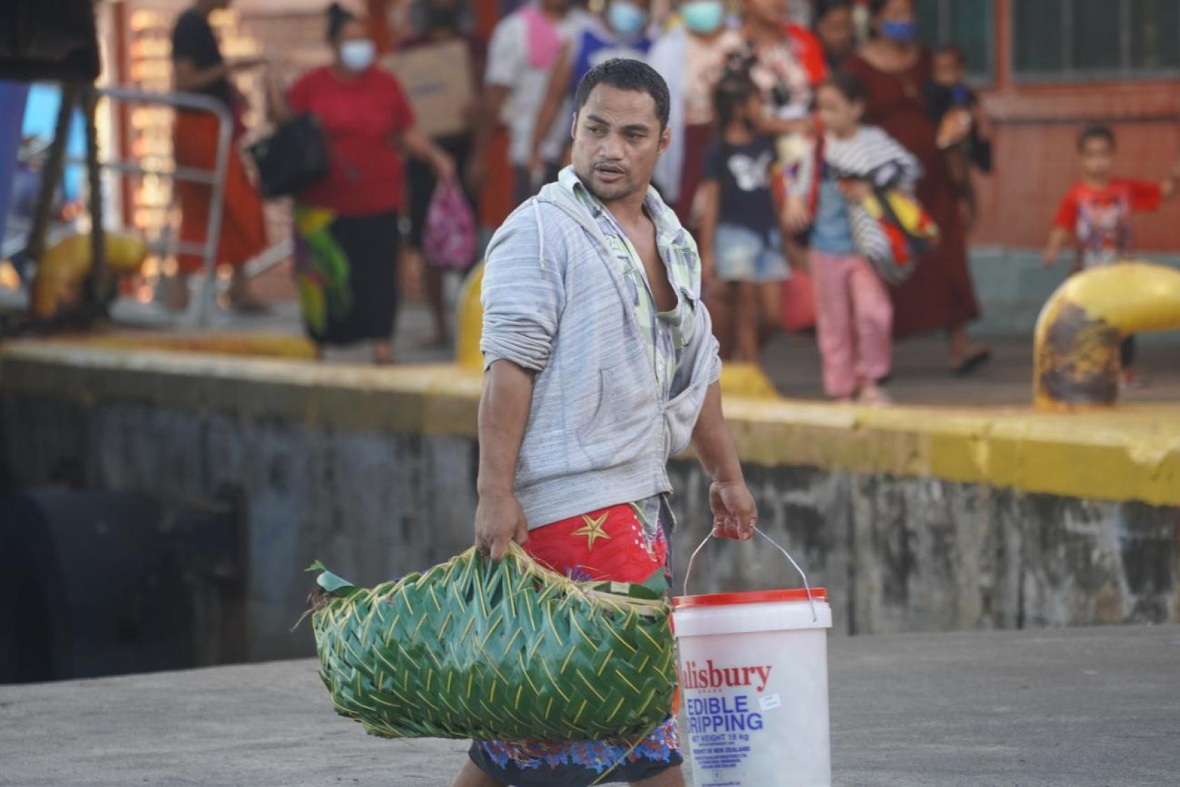 A person is walking in a busy street market carrying a large green leaf-woven basket on their left side and a white plastic bucket with red text on the right
