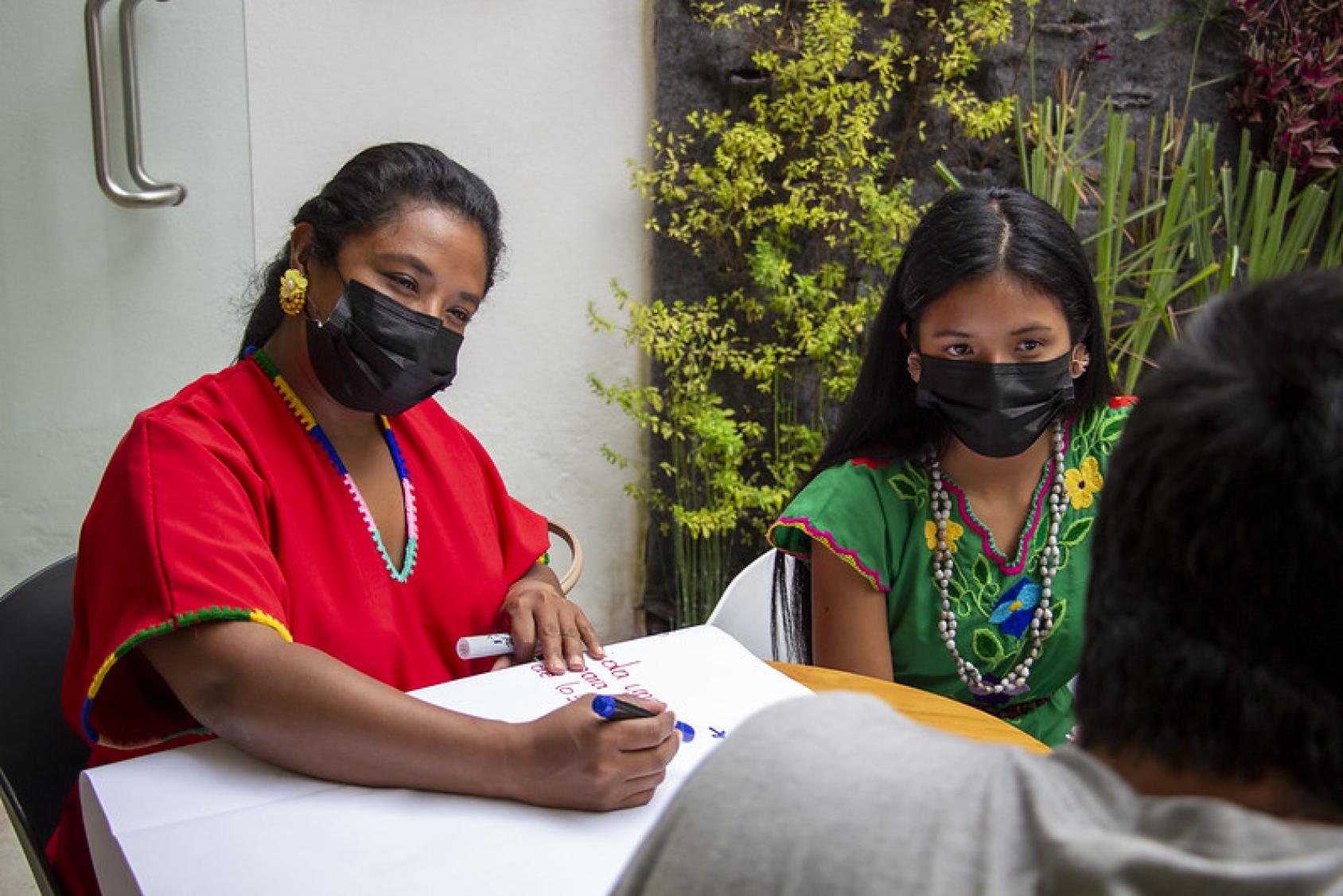 Two women sit at a table and write on a large white board, they are wearing masks but looking at the camera and smiling 