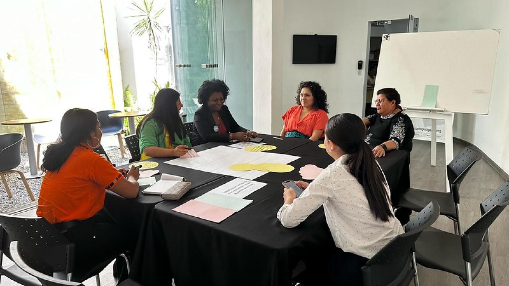 A group of women from diverse backgrounds sit at a table together facing each other and talking. They have notepaper in front of them and are working on something