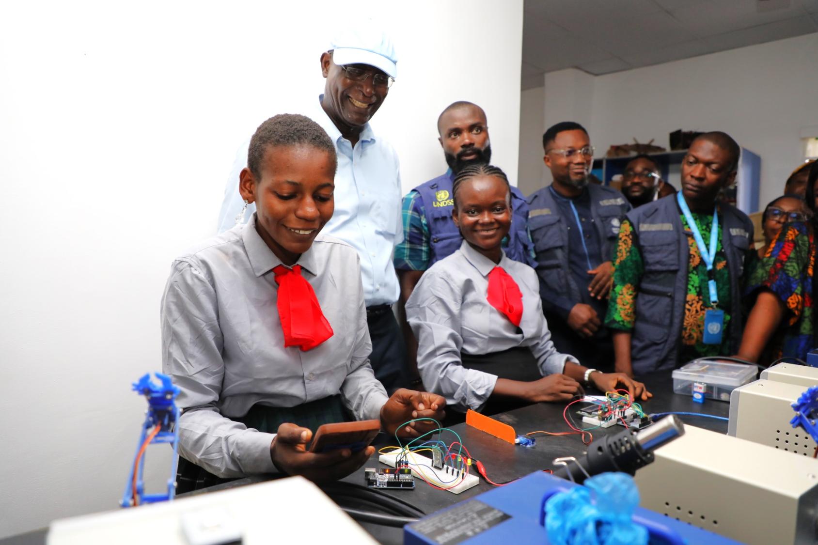 Two female students using electronic devices, while a group of adults looks on