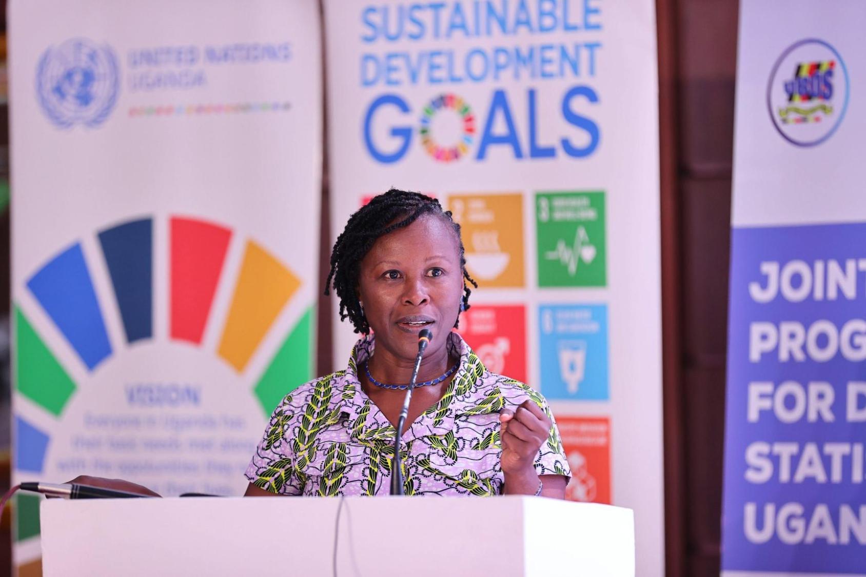 A woman wearing a green and white dress stands at a podium with the SDG banners behind her