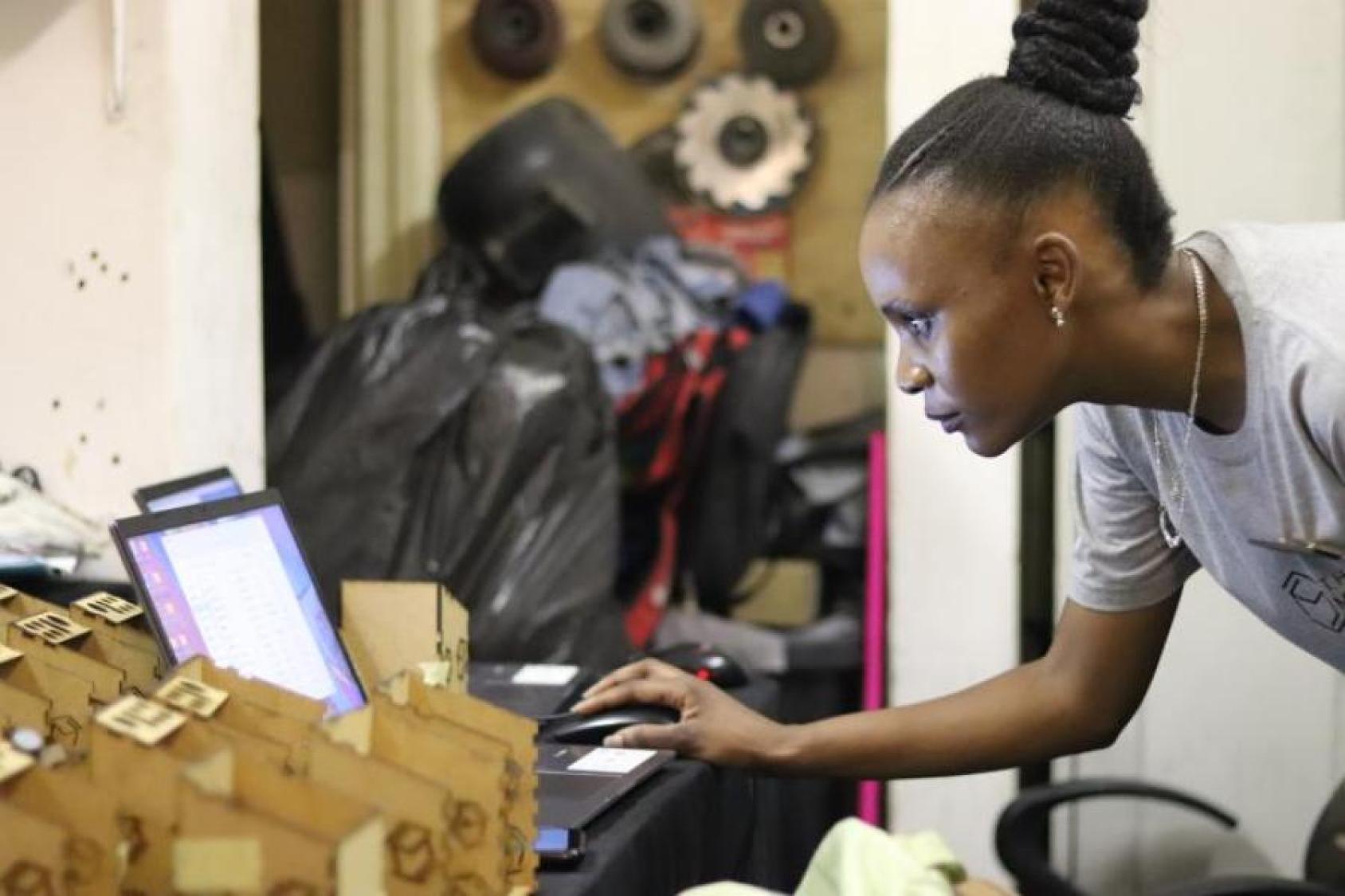 A young woman in front of a computer with a busy workshop background behind her