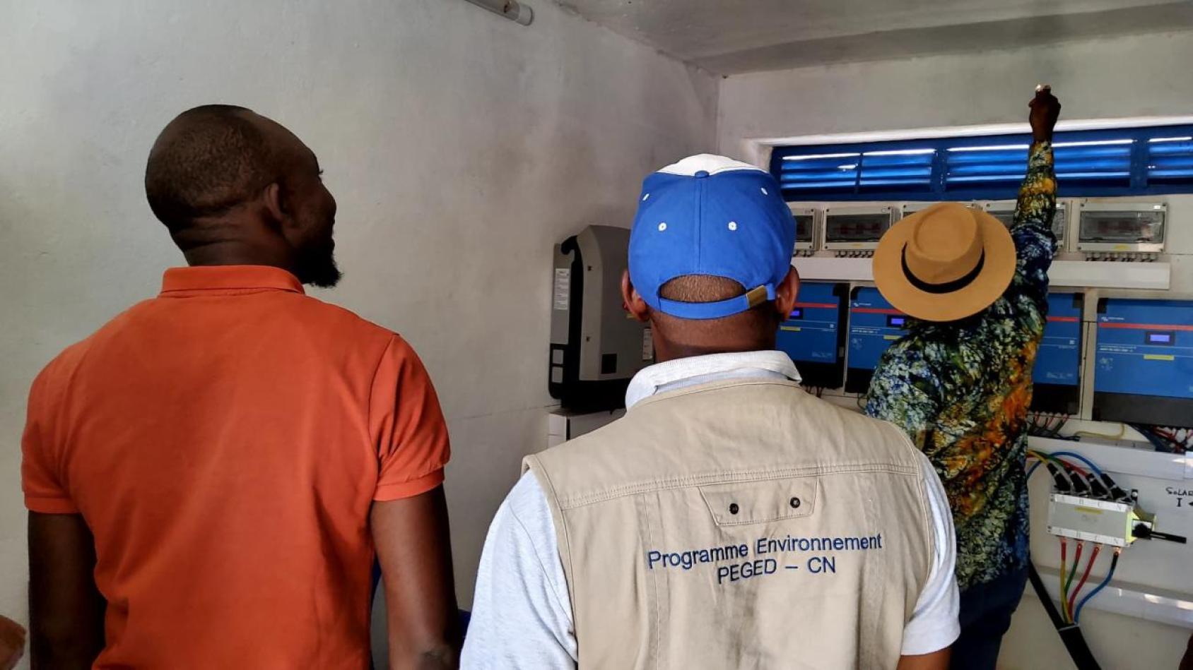 A group of people stand in front of a switch board. One of them is wearing a vest that says UNDP Guinea (in French)