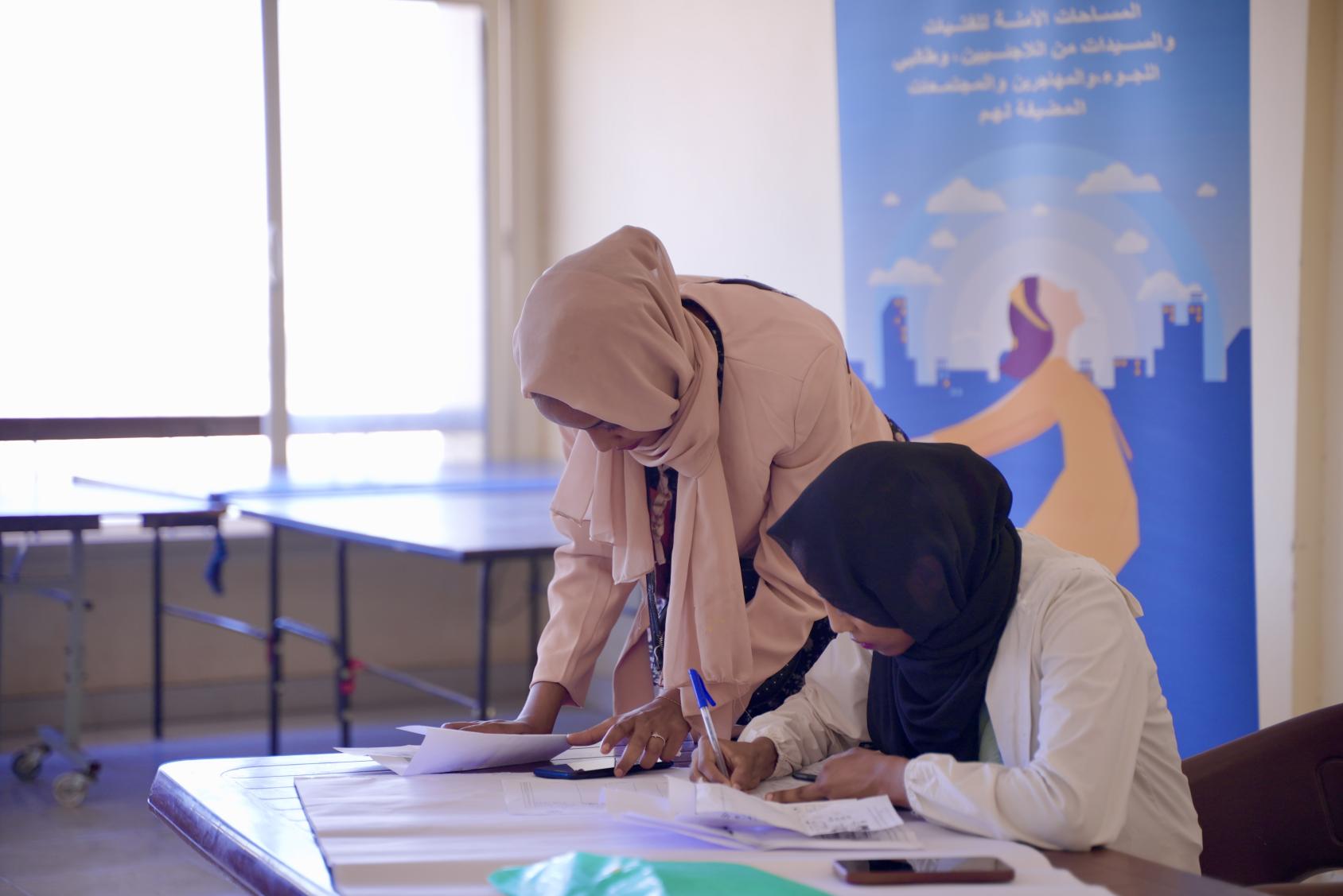 Two women are taking notes and behind them is a banner that discusses programmes on supporting women report and combat violence against women