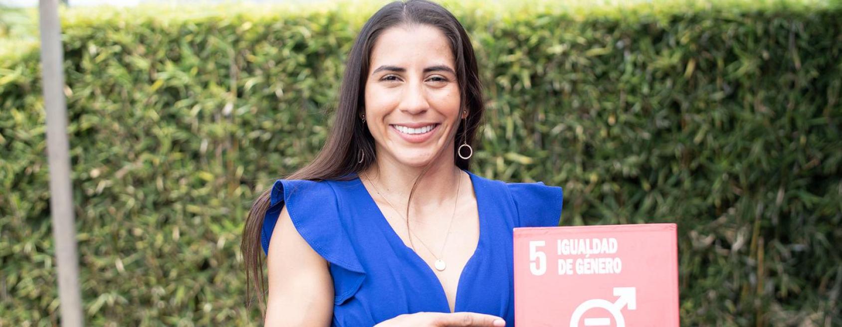 A woman is wearing a blue dress and holding a box that reads Gender Equality. She is a champion in speaking up against Gender-Based Violence