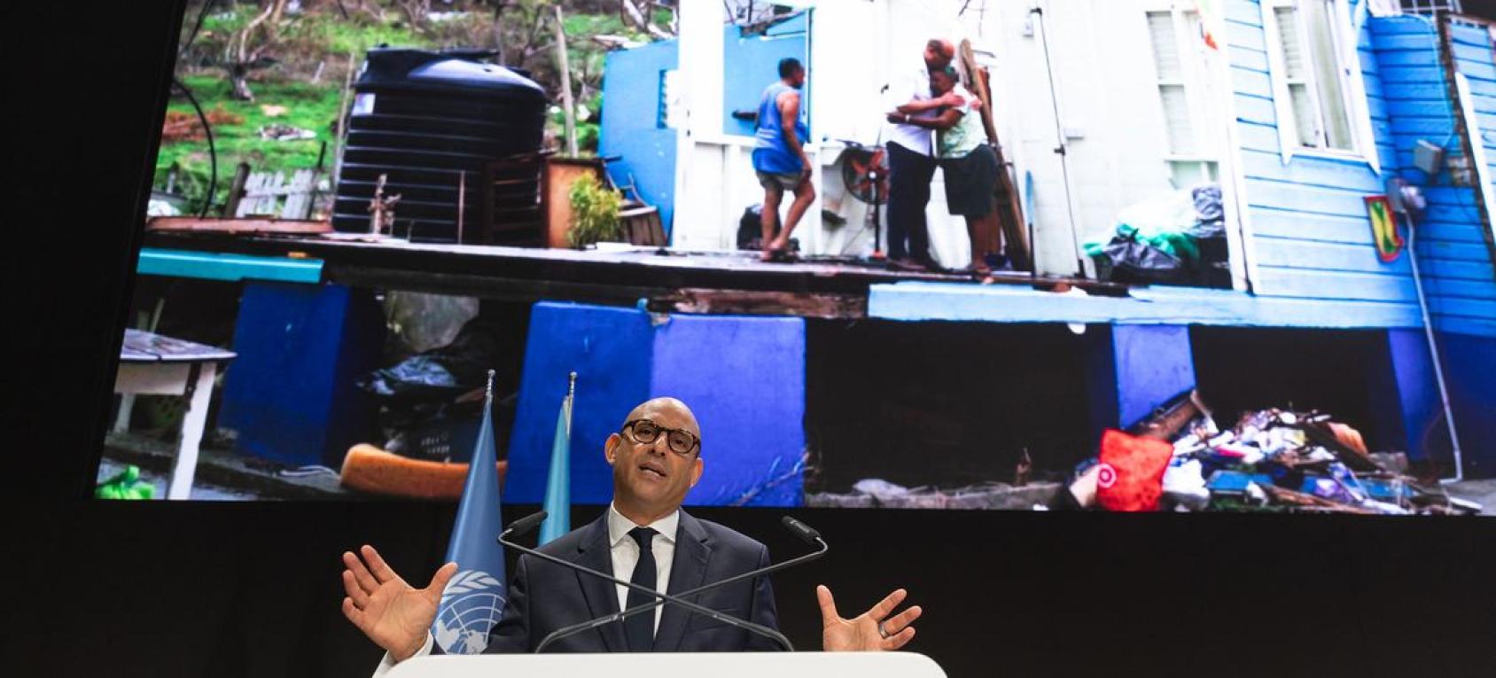 A man standing in front of a devastating scene on a screen and speaking at a podium