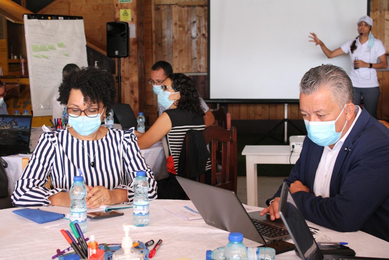 Several people look at documents and computers while a person speaks in the background.