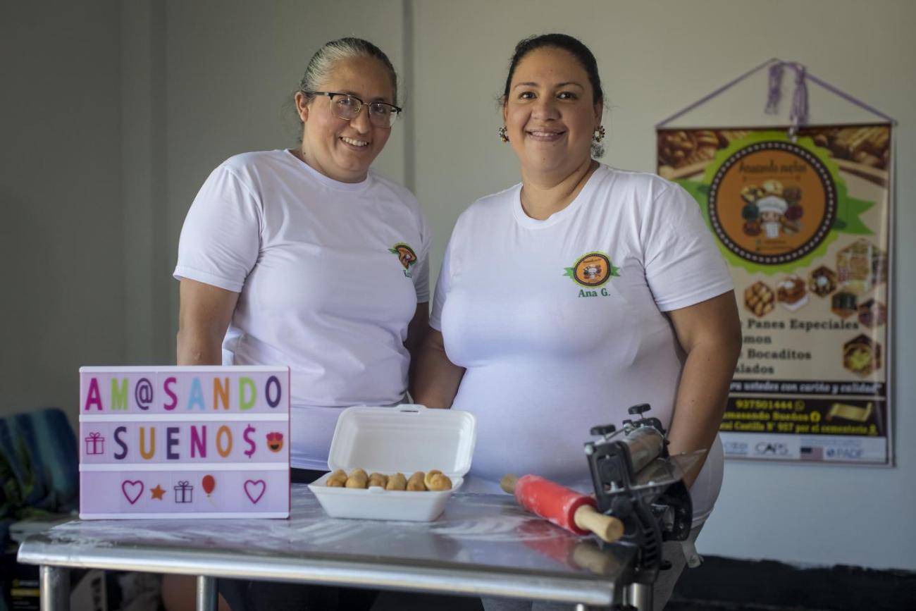 two women in white t-shirts stand behind a kitchen counter 