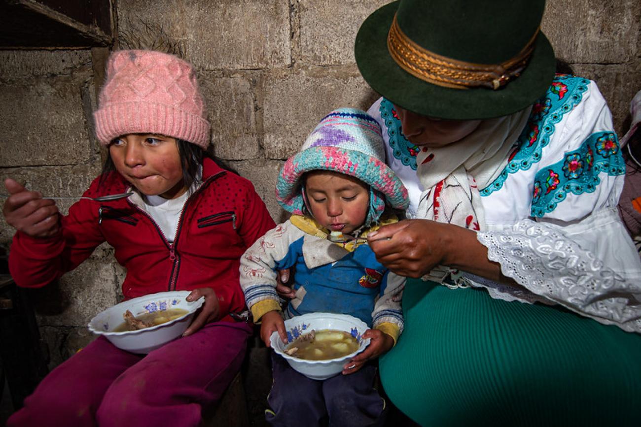 two young children eat a bowel of soup next to their mother 