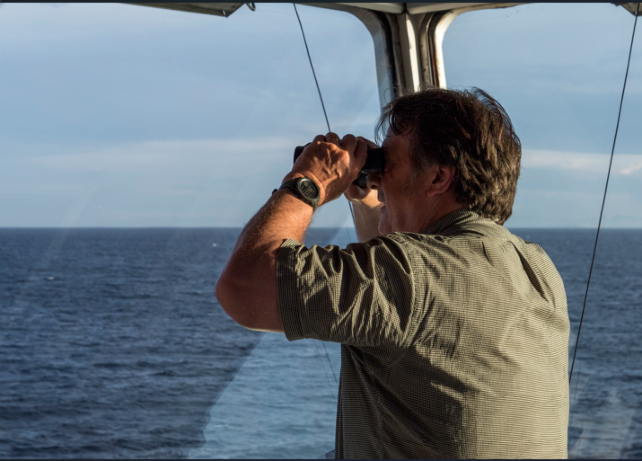 A man in a grey shirt aboard a ship looks out into the sea with binoculars