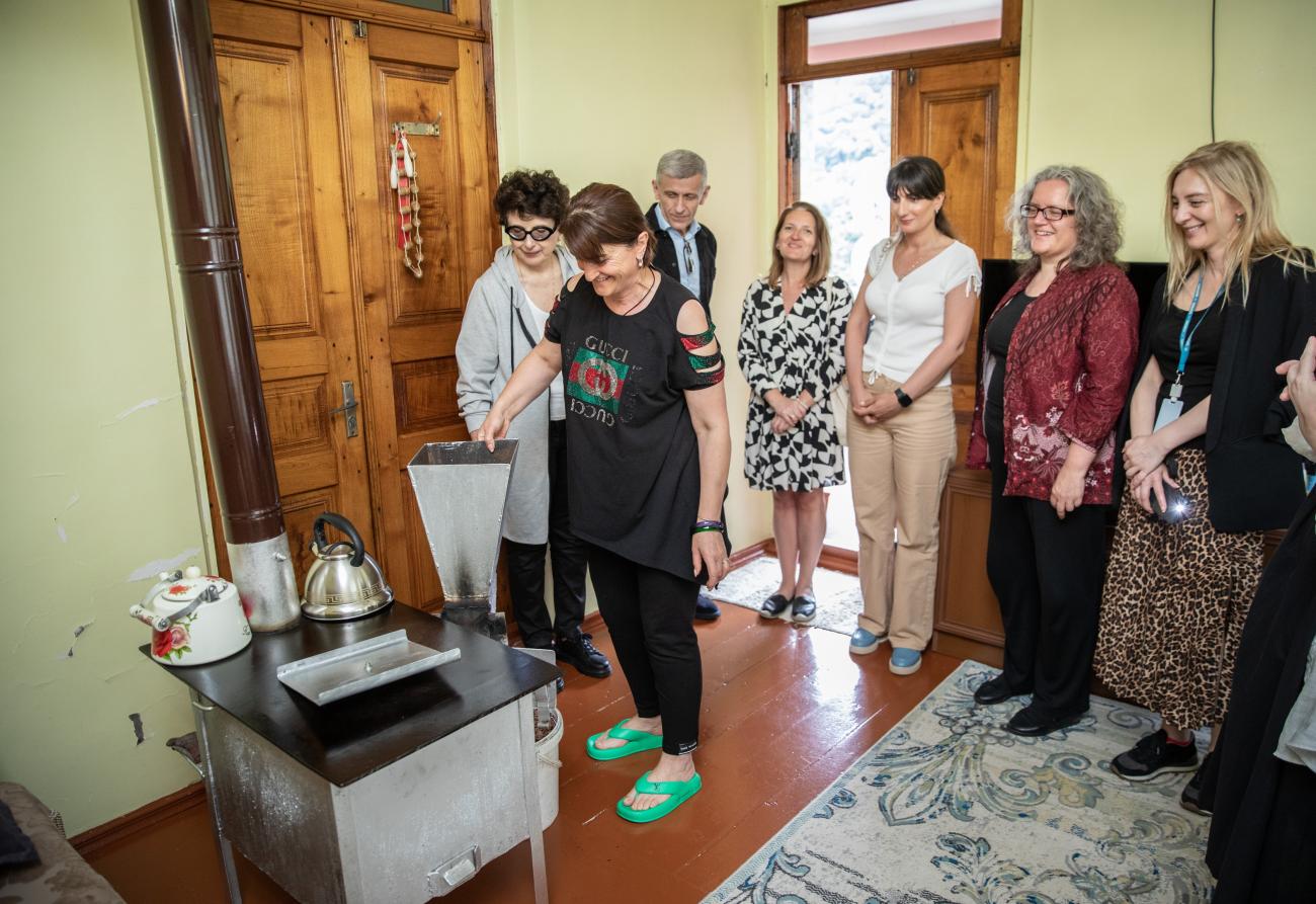 group of people gather in a circle in someone's home to watch a woman use a renewable energy stove.