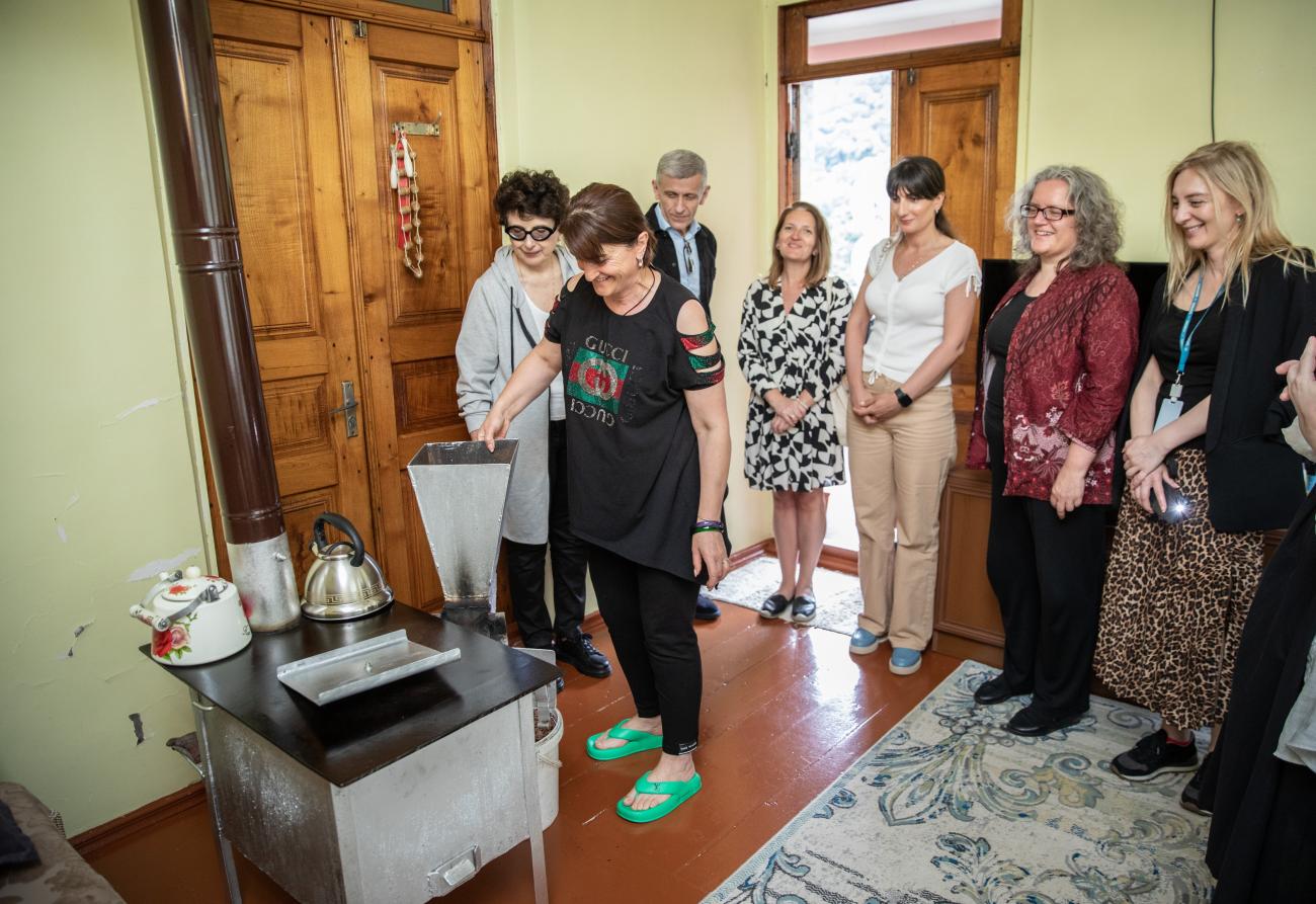 group of people gather in a circle in someone's home to watch a woman use a renewable energy stove. 