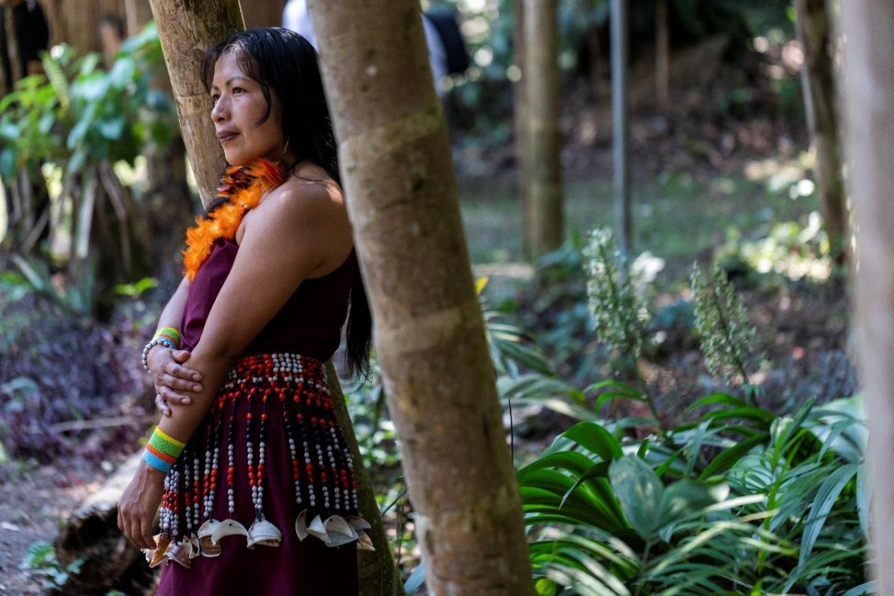 woman with dark hard and traditional indigenous dress stands side ways against a tree 