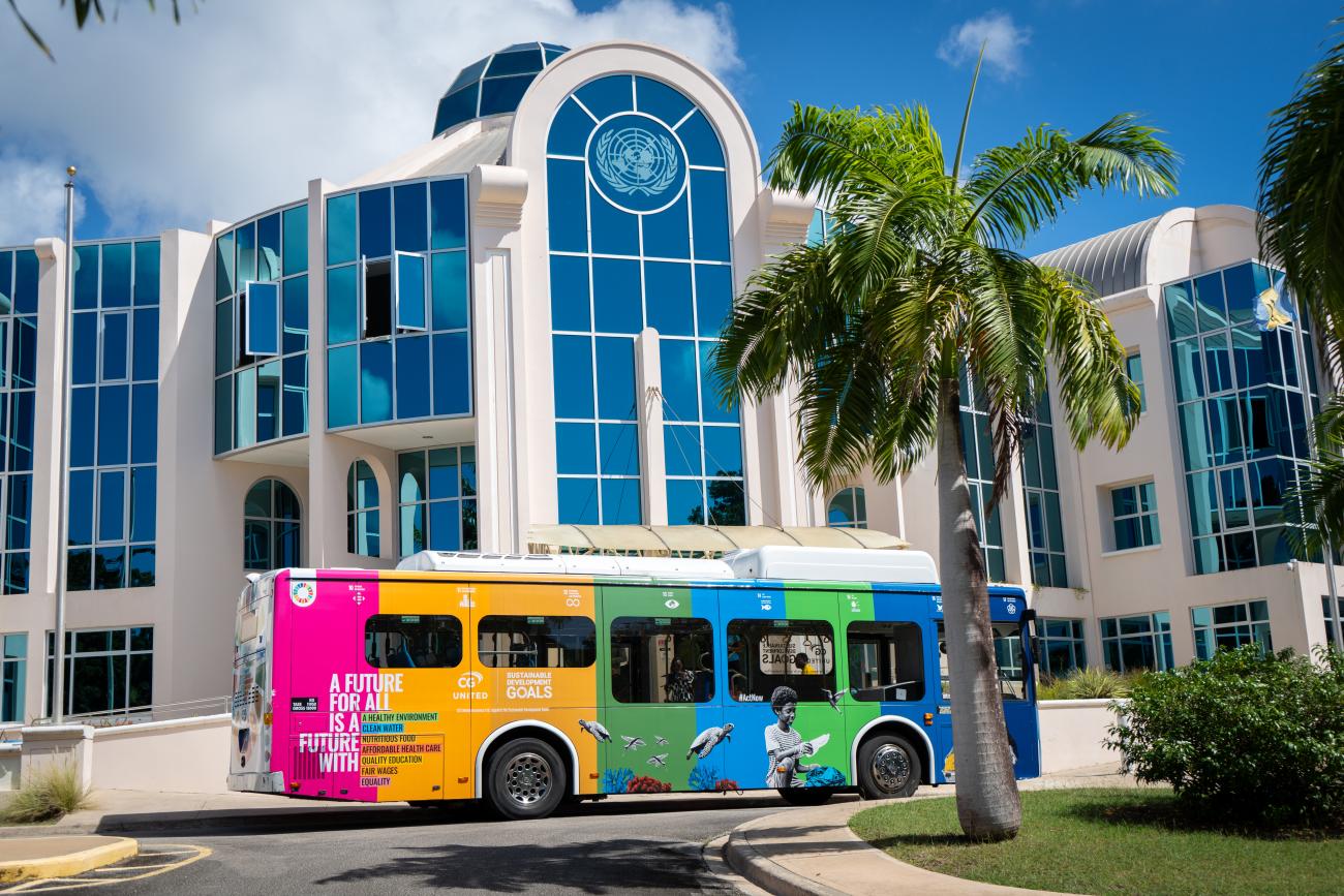 A multi-coloured bus standing in front a tall blue building (the UN in Barbados).