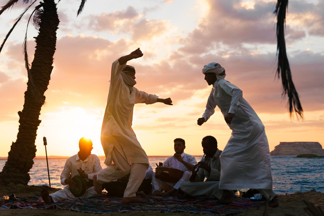 A group of men in white clothing dance together by a beachside as the sun sets in the background