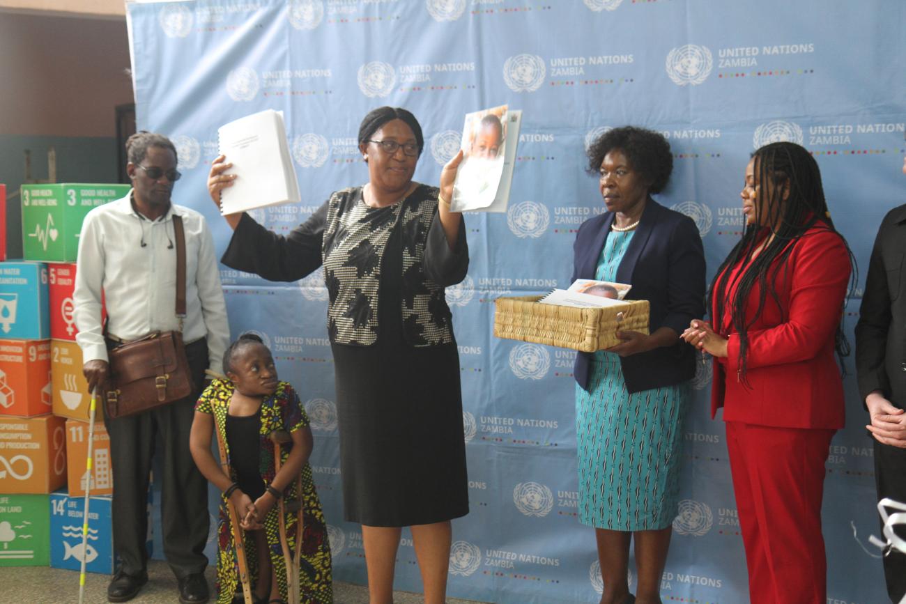 A group of five individuals standing in front of a backdrop that has the United Nations logo and some text, along with logos representing different Sustainable Development Goals. The individuals are holding various items including papers and a basket.