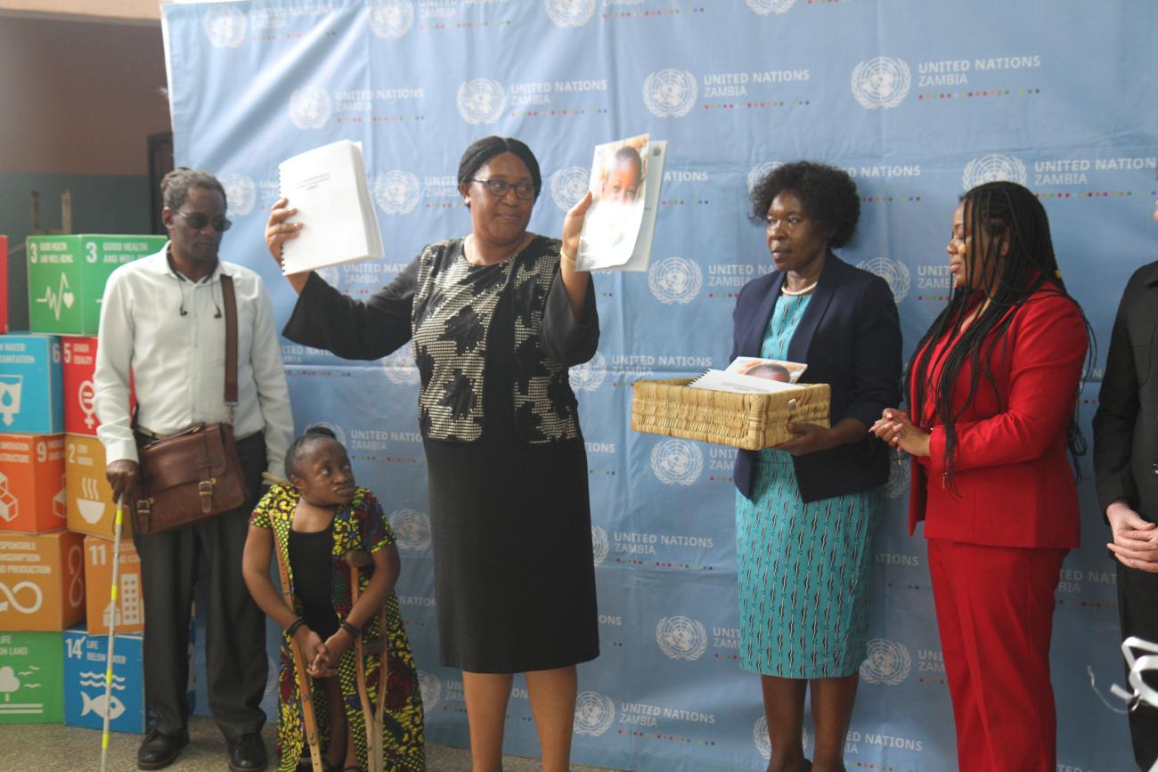 A group of five individuals standing in front of a backdrop that has the United Nations logo and some text, along with logos representing different Sustainable Development Goals. The individuals are holding various items including papers and a basket. 