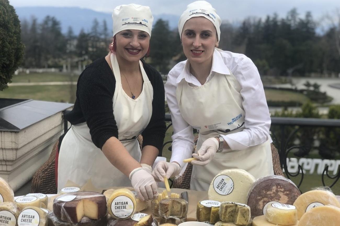 Two individuals in aprons and chef hats arranging a variety of artisan cheeses on a table outdoors