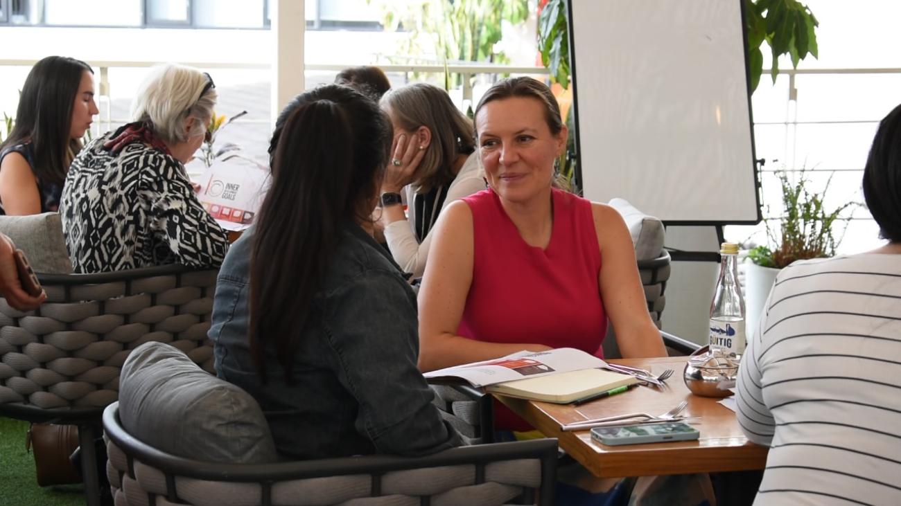 A woman in a red shirt talkng to another woman in a black shirt, there are other women in the background sitting on chairs and talking