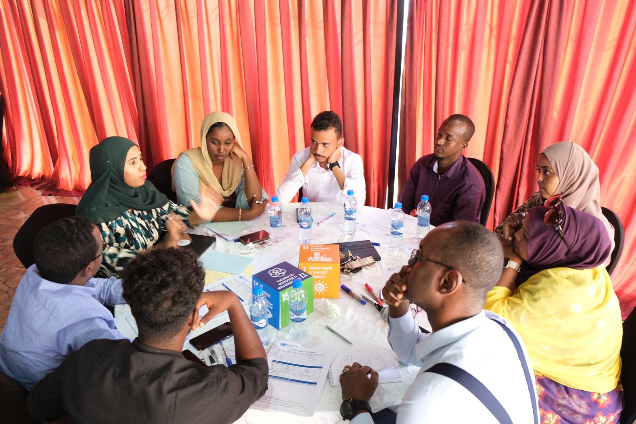 A group of four women and five men gather around a table, in discussion. Papers are on the table along with blocks for the SDG Goals.