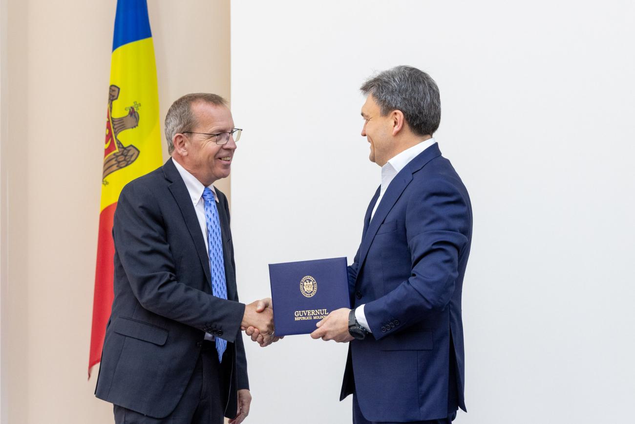 Two men in dark suits exchanging a document, shaking hands in front of a Moldovan flag. 