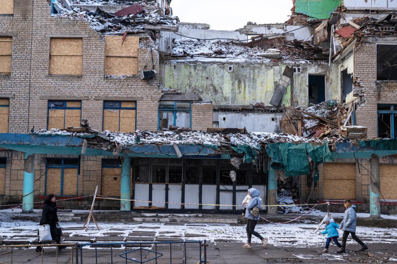 The image depicts a severely damaged multi-story building with its upper part largely destroyed, exposing the interior rooms. In the foreground, three people, including a child, walk past the building on snow-covered ground. 