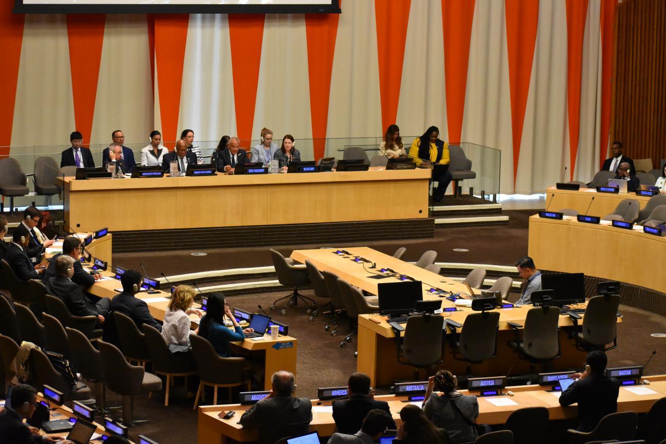 A professional conference room setting with individuals seated at a long table on a stage, facing an audience. Each person at the table has a microphone and papers in front of them, against a backdrop of orange and white vertical stripes. The audience, seated in rows with attached desks and laptops, is attentively engaged in the presentation or discussion taking place.