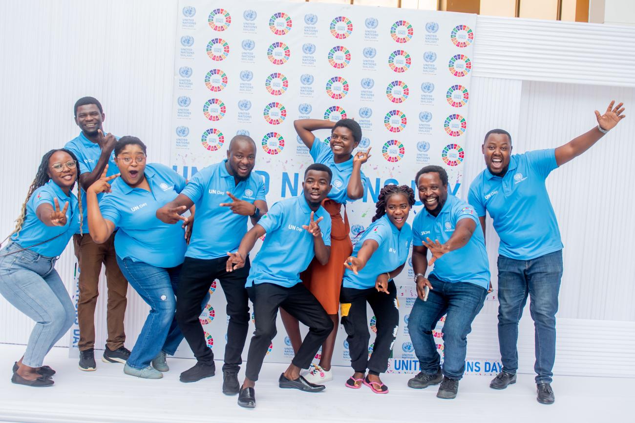 A group of nine young men and women in blue T shirts pose enthusiastically in front of a colourful background