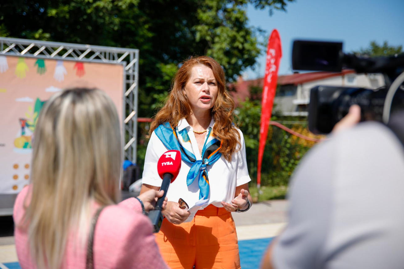 a woman speaks to the media outdoors. There is a camera and microphone pointed at her