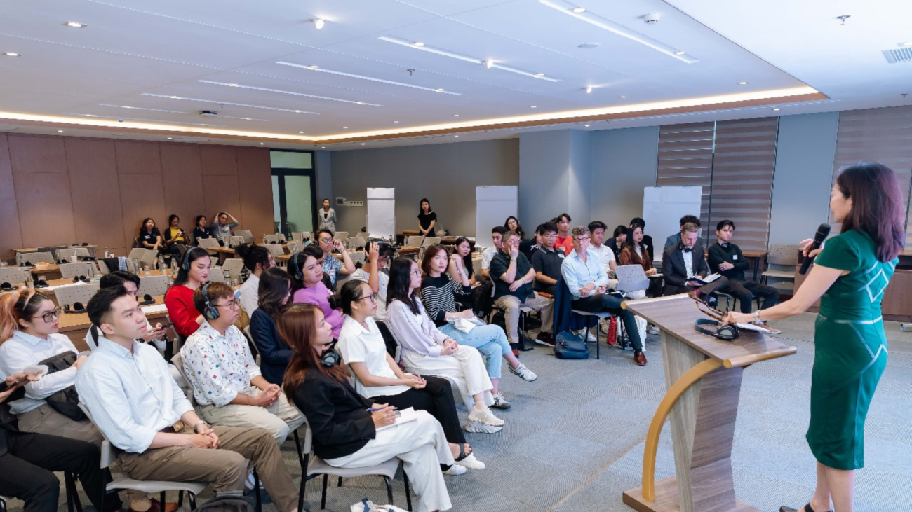 A woman is standing at the lectern and speaking to a group of young people in a conference room