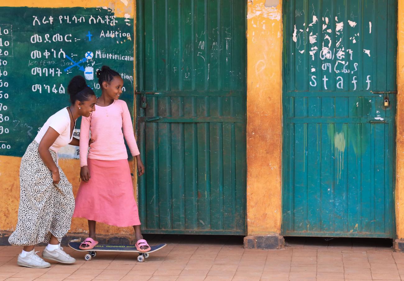 Two girls in front of a classroom, one of them is on a skateboard.