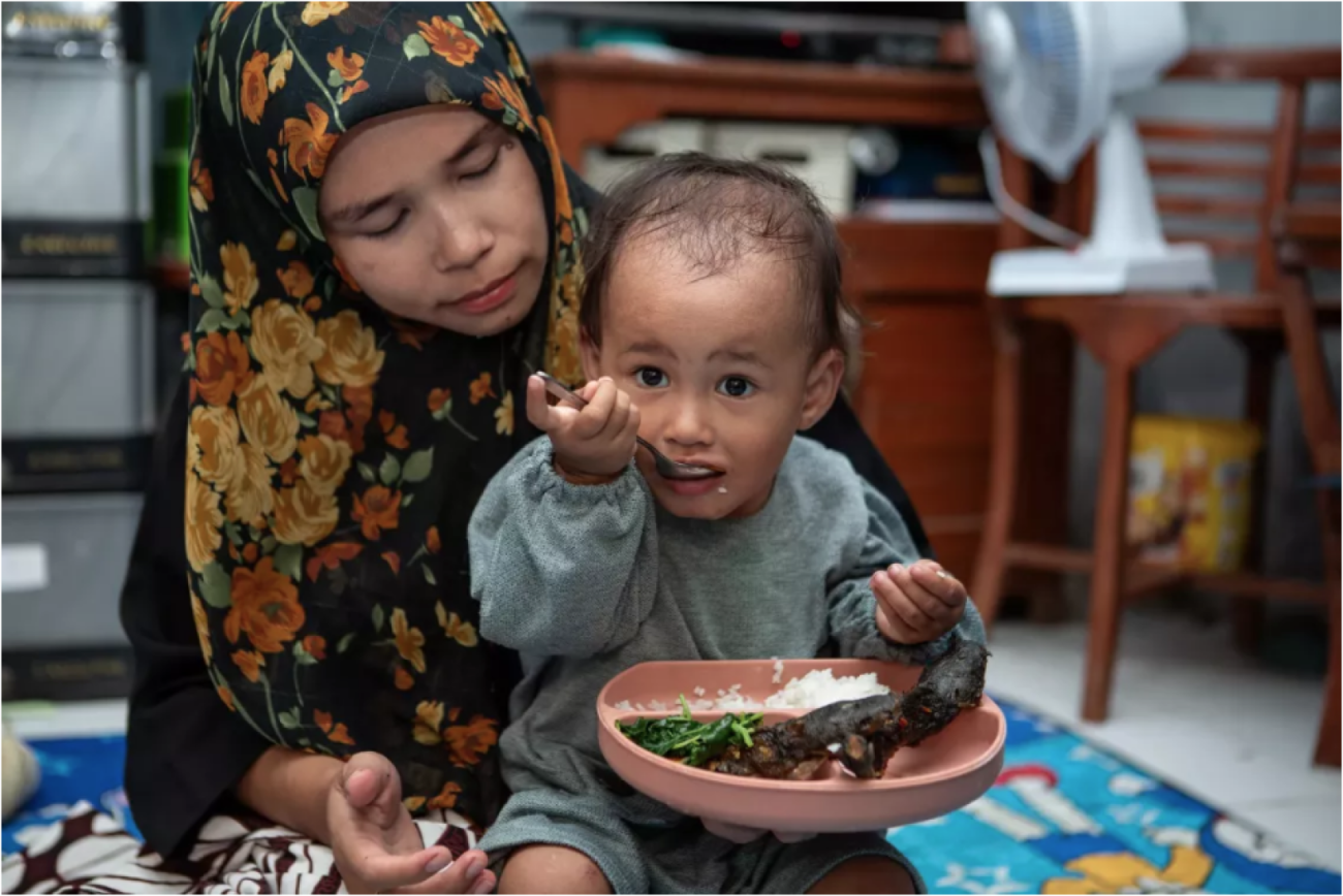 A little boy is eating his food seated on his mother's lap