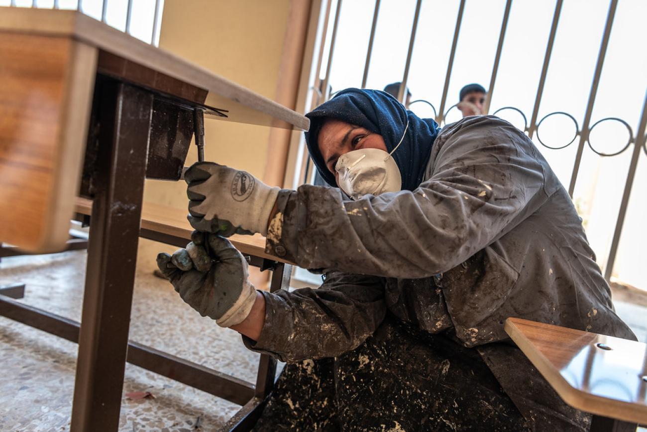 A woman in Iraq refurbishes a desk in a classroom.
