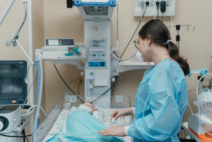 A woman is wearing a blue surgical cape and looking at her baby in an incubator. They are in a room with lots of medical equipment 