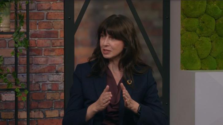 A woman is seated in front of a brickwall background and speaking. She is wearing a black blazer and red top