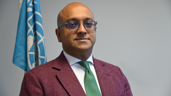 The RC is standing in front of a grey background with the UN flag behind him. He is wearing a red jacket and glasses