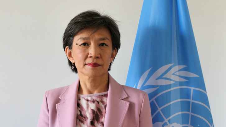 The RC is wearing a pink jacket and standing in front of a grey background with the UN flag