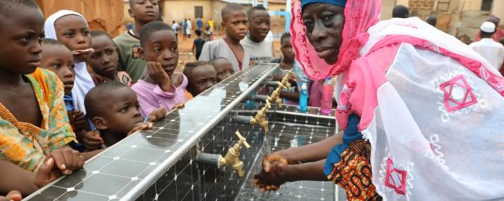 A group of children and a woman gather around water faucets with running water