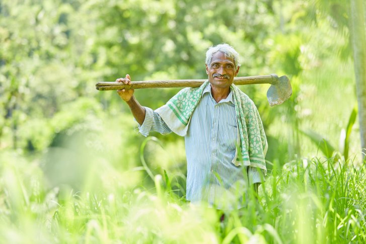 A smiling farmer carries a hoe on his shoulders while standing in a green field.