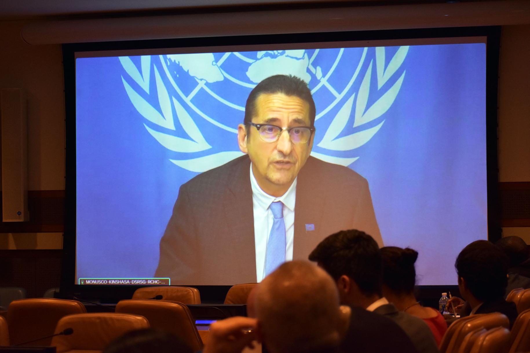 man in dark suit speaks to meeting on screen with UN flag behind 